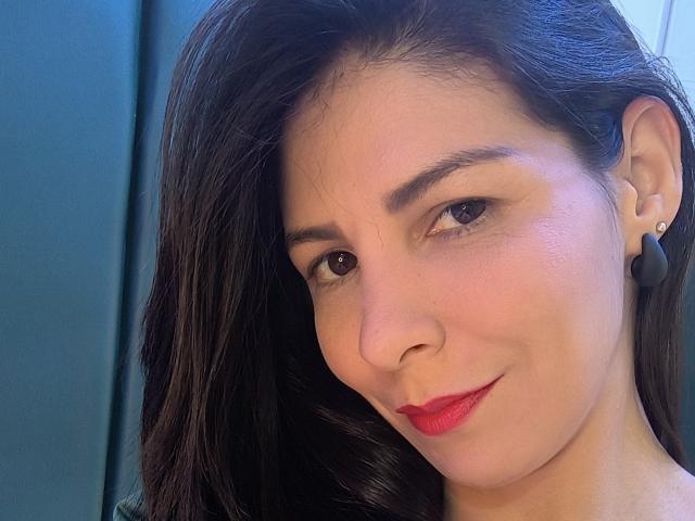 Il viso di EliseBell, capelli castano scuro ondulati, rossetto rosso, leggero sorriso, occhi marroni, pelle chiara, piccoli orecchini neri, luce soffusa, testa leggermente inclinata, trucco leggero, carina..