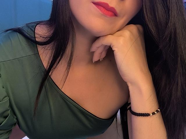 EliseBell, avec ses cheveux foncés, son t-shirt vert et son rouge à lèvres rouge, appuyant son menton sur sa main, portant un bracelet de perles noires, montrant ses seins..