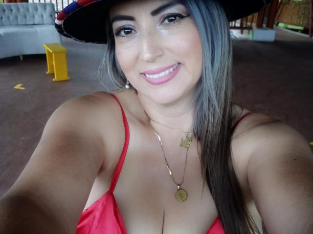 EvaNathaly with gray hair, wearing a red top, showing her big tits, smiling, wearing a hat, gold necklace, and pink lipstick, in a selfie.