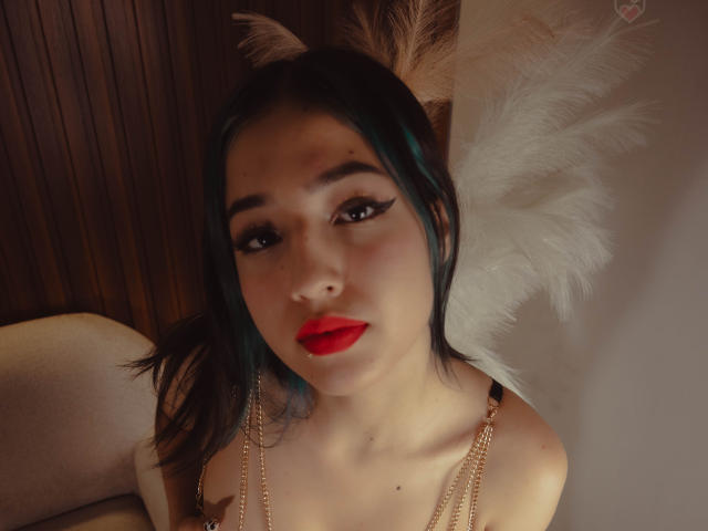 LizzyQueenmml hat blasse Haut, dunkelgrünes Haar, roten Lippenstift und trägt weiße Federflügel mit goldenen Ketten. Sie blickt mit verführerischen, schwarz umrandeten Augen in die Kamera..