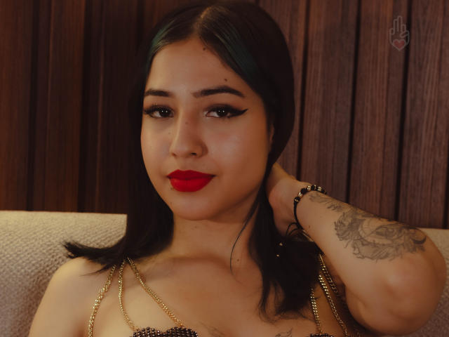 LizzyQueenmml hat dunkelgrüne Zöpfe, roten Lippenstift, schwarzen Eyeliner, eine goldene Kette um den Hals, einen tätowierten Arm und blickt mit einem leichten Lächeln in die Kamera. Brüste sind sichtbar..