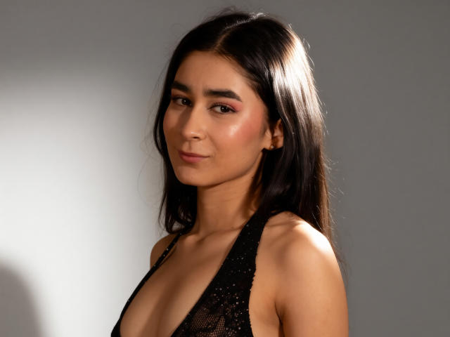 RachellFab, avec ses cheveux foncés, son fard à paupières rose et son haut en dentelle noire qui dévoile ses seins, regarde l'appareil photo avec un léger sourire. Sa peau est bronzée et elle est séduisante..