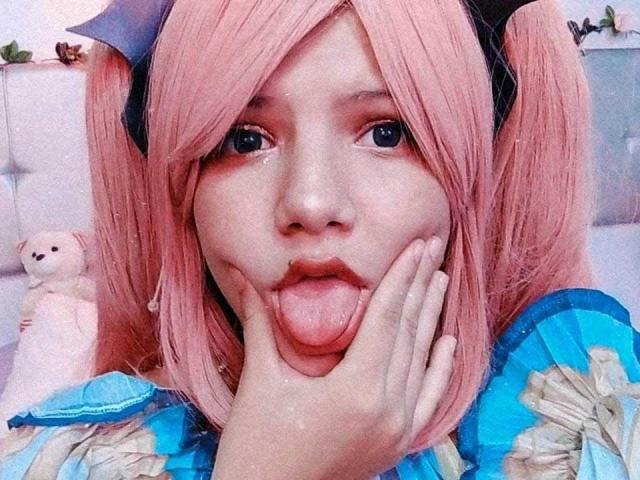 MiaBellee with pink pigtails and blue frilled top, sticking out tongue, hand on cheek, blue eyes, cute face, black bow, plush toy in background, soft lighting.