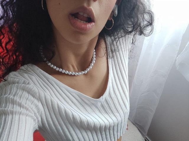CurlyBlossom's face, showing parted pink lips, with curly dark hair, wearing a white ribbed top, pearl necklace, and gold hoop earrings, in a bright room.