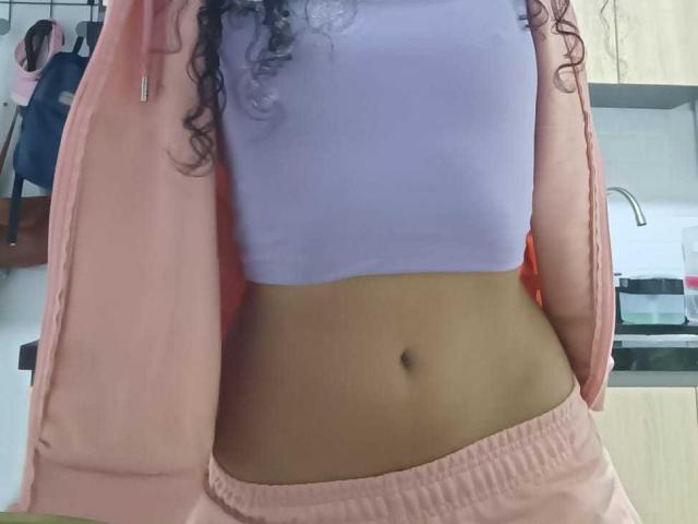CurlyBlossom has a purple crop top, showing her midriff with a navel, and is wearing pink pants with a pink jacket open. Her curly hair is visible.