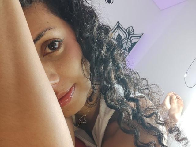 CurlyBlossom with curly black hair, brown eyes, and red lips, lying down, arms raised, wearing a star necklace, legs in the air, smiling slightly, in a relaxed pose.