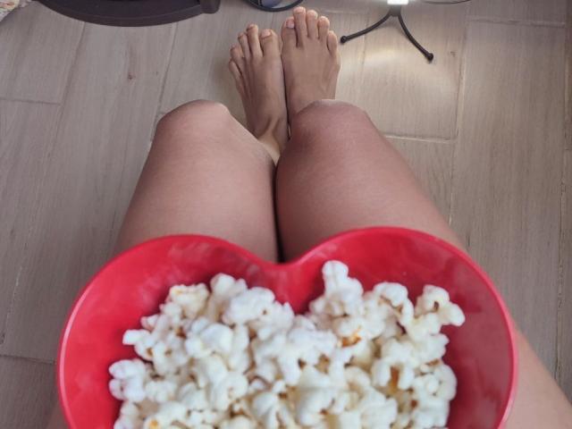 CurlyBlossom's legs with a heart-shaped red bowl of popcorn in front of them, bare feet on wooden floor, from a top-down perspective.