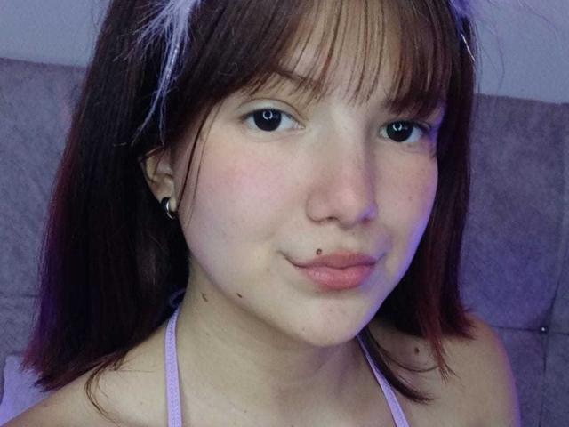 MiaBellee has light brown hair with bangs, small dark eyes, a nose piercing, and a lip piercing. She's wearing a pink halter top, looking at the camera.