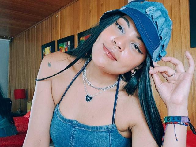 AntonellaFioree with dark hair, blue cap, denim halter top, nose ring, chain necklace, small star tattoo, smiling, touching hat, red bracelet, wood-paneled wall.