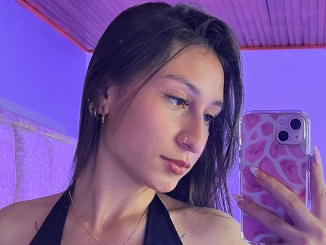 MaddyCarina, con el cabello castaño mojado, un piercing en la nariz y lápiz labial rosa, sosteniendo una funda rosa para teléfono con estampado de leopardo, mostrando sus pechos con un top negro de tirantes, bajo una luz púrpura..