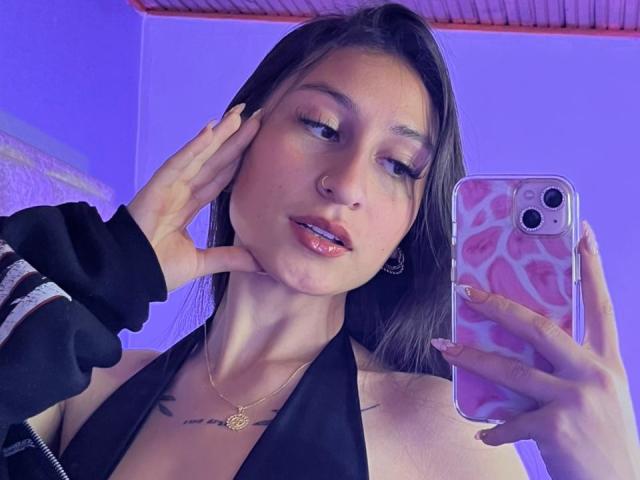 MaddyCarina with long brown hair, wearing a black halter top, holding a pink phone with leopard print case, touching her cheek, nose ring, and gold necklace. Her tits are visible.