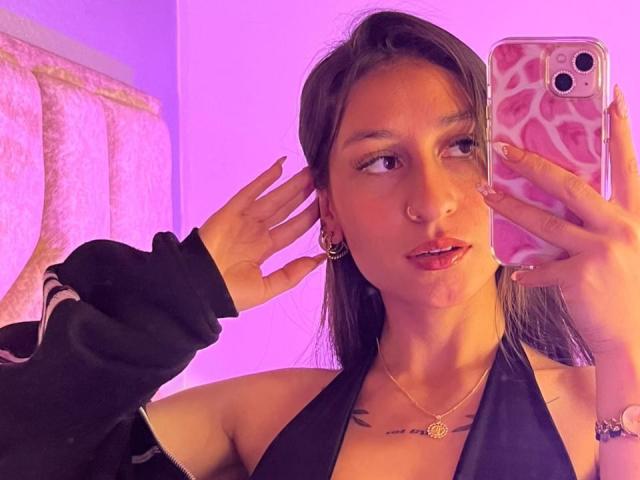 MaddyCarina with dark hair, nose ring, and pink phone case, holding phone, black halter top, purple lighting, black jacket, gold necklace, and earrings, taking a selfie.