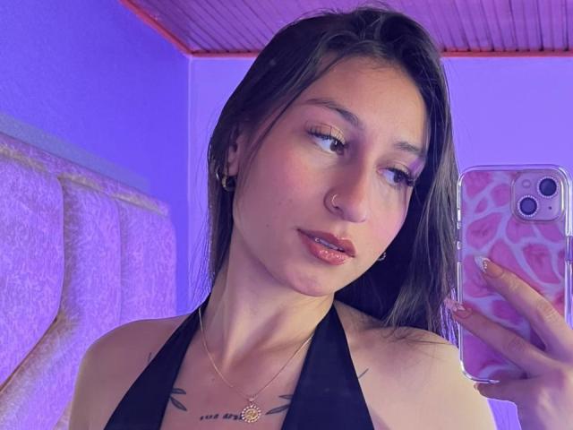 MaddyCarina with black hair, nose ring, and pink lipstick, holding a pink phone with leopard print case, wearing a black halter top, showing small tits and a tattoo.