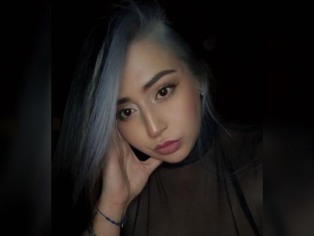 SaraDeLuna, avec ses cheveux bleu pâle, ses yeux marron et ses lèvres roses, portant un haut transparent, la tête reposant sur sa main, regardant légèrement vers le bas, dans un environnement sombre..