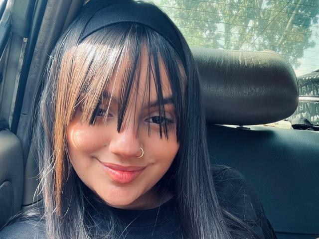 ValentinnaSexx has black hair with blonde highlights, a nose ring, and a black headband, smiling in a car with a black leather headrest. She's wearing a black shirt.
