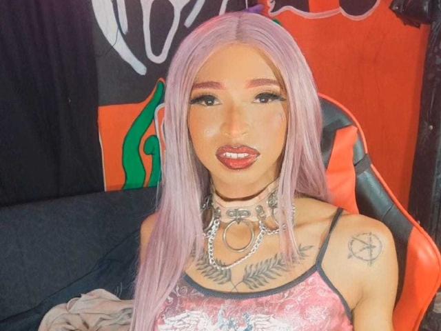 EveDollyx, con lunghi capelli rosa, rossetto rosso e tatuaggi, indossa una collana a catena e una camicetta di pizzo rosa, seduta su una sedia arancione, che mostra il sedere e il seno..