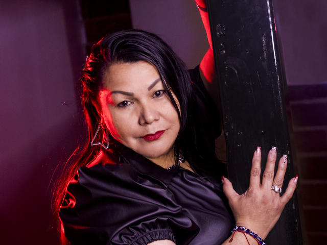EllenaBoobs with black hair, red lipstick, and a black satin top, gripping a pole, looking at the camera with a slight smile, red light highlighting her face.
