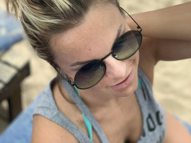 BelleLisaG with blonde hair in a bun, wearing dark sunglasses and a gray tank top, showing her tits and a blue strap, smiling slightly at the camera.