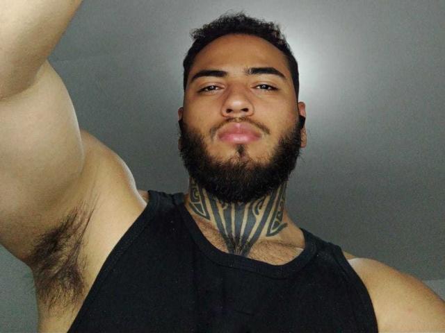 AlbertAmin69, een gespierde man met donker krullend haar en baard, een zwart tanktop, een grote tribale tatoeage op zijn nek, okselhaar, een serieuze uitdrukking, die een selfie maakt vanuit een lage hoek..