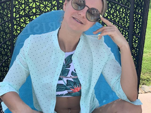 BelleLisaG with sunglasses, light green polka-dot shirt, tropical patterned bikini top, blue towel, and green nails, sitting outside, smiling, right hand near face.