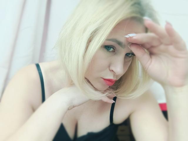 MagraLi con un caschetto biondo, labbra rosse, biancheria intima nera e unghie bianche, appoggia il mento sulla mano, guardando seducentemente la fotocamera. Il seno è visibile attraverso lo spallino nero..