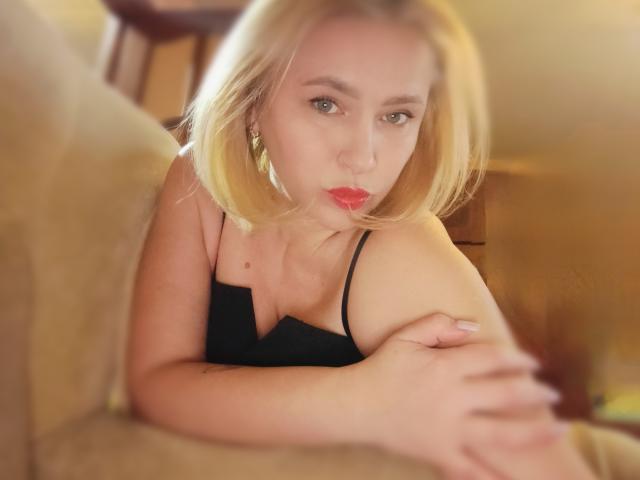 MagraLi with blonde bob, pouty red lips, black tank top, looking at camera, arms resting on beige couch, soft focus, slightly blurry, showing her tits and cute face.