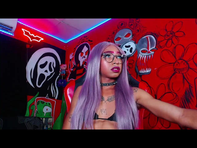 EveDollyx, avec ses longs cheveux violets, ses lunettes et son rouge à lèvres rose, exhibe sa petite poitrine dans un soutien-gorge noir, portant un collier, assise dans une pièce éclairée au néon et décorée de crânes..