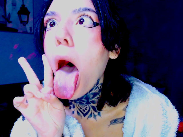 Lannie Gonzales, con cabello negro, ojos grandes y la lengua afuera, haciendo un gesto de "rock and roll", mostrando el cuello tatuado, y vistiendo una blusa blanca y esponjosa. Su lengua es rosa y húmeda..