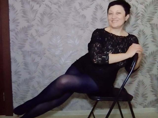 EmilyLlove with black lace top and dark tights, sitting on a chair with one leg stretched out, smiling, hands on the chair, short dark hair, fair skin.