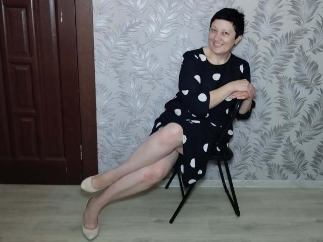 EmilyLlove sits on a black chair, wearing a black dress with white polka dots, beige heels, and short black hair, smiling with crossed legs and arms.