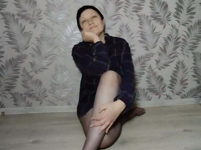 EmilyLlove is sitting on the floor with one leg up, wearing a dark plaid shirt and sheer black tights, resting her chin on her hand, looking up.