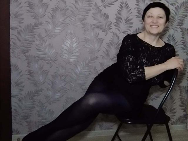 EmilyLlove with short black hair, smiling, wearing a black lace top and black tights, sitting on a black chair, legs spread, showing her taut ass.