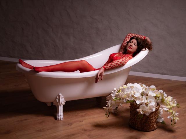 RoseHille in red fishnet lingerie and tights, reclining in a white clawfoot bathtub, wearing a red hat, with white flowers in a wicker basket on wooden floor.