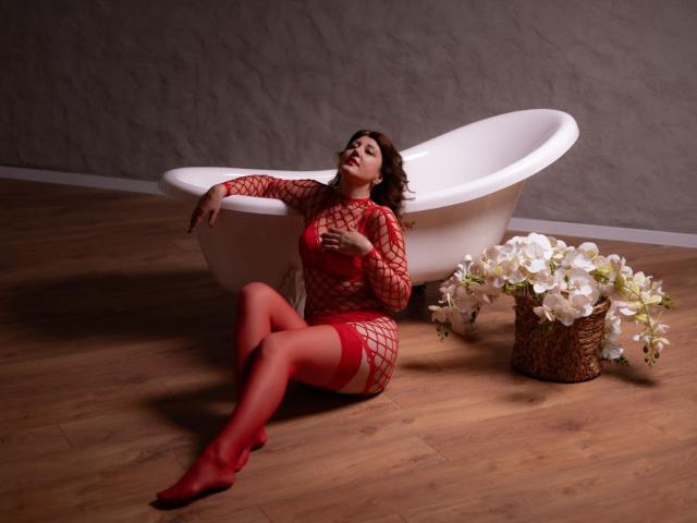 RoseHille sits on wooden floor wearing red fishnet lingerie and red stockings, arms resting on white bathtub, legs crossed, with wicker vase of white flowers to the right.