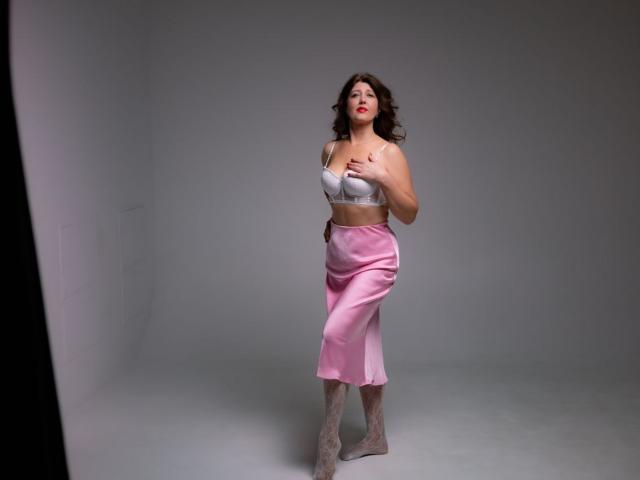 RoseHille stands in a gray studio, wearing a silver bra, pink satin skirt, and lace boots, with curly brown hair and red lipstick, touching her chest.