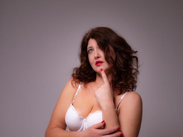 RoseHille, a fair-skinned woman with wavy brown hair, stands against a plain gray background. She wears a white lace bra with silver straps, her left hand resting on her chest while her right index finger touches her chin. She has red lipstick and red nail polish, her expression thoughtful. The composition is centered, highlighting her upper body and facial features. The image uses soft lighting to emphasize her curly hair and the texture of her bra.