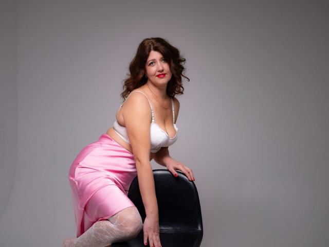 RoseHille with curly brown hair, white bra, pink satin skirt, and white lace stockings, leaning on a black chair, showing her curvy ass and medium-sized tits.