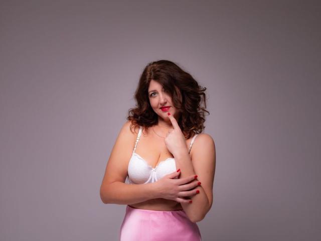 RoseHille stands against a plain gray background, wearing a white lace bra that accentuates her medium-sized breasts. She has curly, dark brown hair and is looking to the side with a slight, confident smile. Her right index finger is raised to her lips, while her left arm crosses over her chest. She's wearing a pink, high-waisted skirt that contrasts with her white bra. RoseHille's red nail polish adds a pop of color. The lighting is even, highlighting her fair skin and soft features. The image has a realistic, studio-style composition.