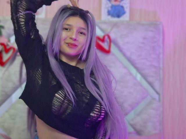 CandySwan with long purple hair, wearing a black, ripped long-sleeve top, showing her tits, arms raised, smiling, in a pink-lit room.