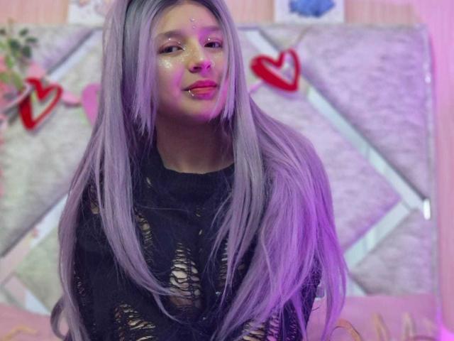 CandySwan with long purple hair, black ripped shirt, glitter on face, pink lipstick, and nose ring, looking up with a slight smile. Pink and purple lighting.
