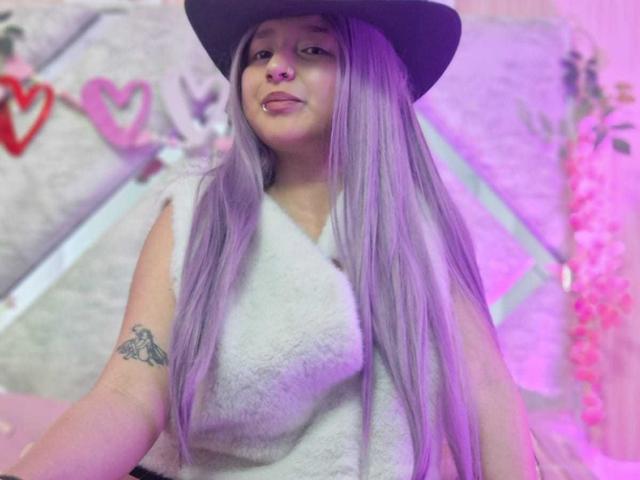 CandySwan with long purple hair, wearing a black hat, white fuzzy top, and nose ring, showing a tattoo on her right arm, smiling slightly.