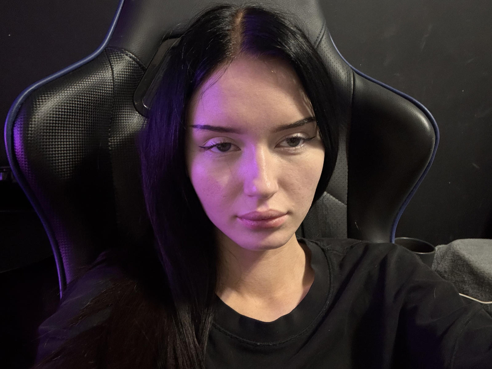NaMacadam, met lang zwart haar, een zwart shirt en een serieuze uitdrukking, zittend in een zwarte gamingstoel, recht voor de camera kijkend, met een subtiel paars licht aan de linkerzijde van zijn gezicht..