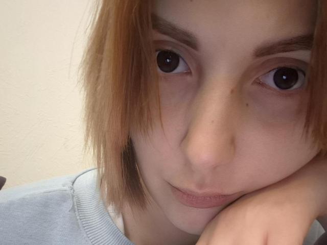 EllaDestiny with light brown hair, brown eyes, and a pensive expression, resting her chin on her hand, wearing a grey shirt, showing her slightly parted lips.