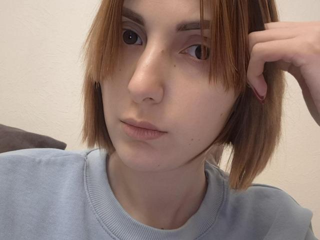 EllaDestiny with shoulder-length brown hair, light skin, and a neutral expression, wearing a gray sweater, touching her right ear with a relaxed hand.