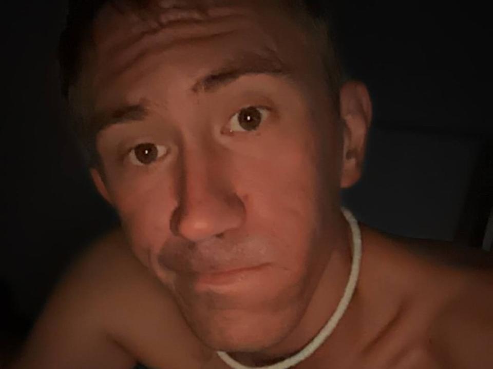 ThiccbtTom è un uomo di mezza età, senza camicia, con capelli castani corti, una leggera barba e una collana bianca, che guarda dritto nella telecamera con un'espressione neutra..