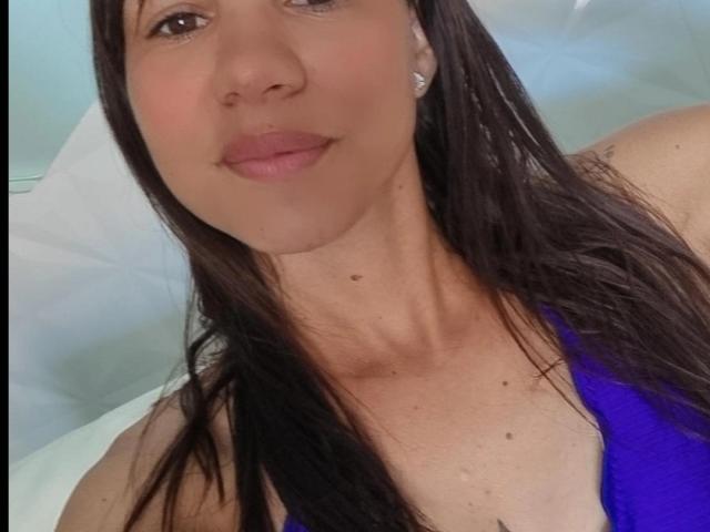 NeztyOne, con cabello castaño, labios rosados y senos pequeños, vestida con una blusa azul, lleva aretes de diamantes, tiene una ligera sonrisa y una pequeña peca en el cuello..