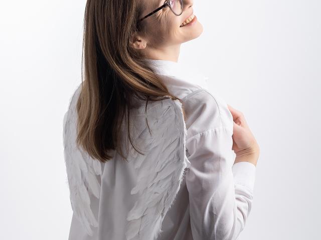 SeaDream stands with her back to the viewer, head turned to the right. She wears a white, long-sleeved shirt with attached angel wings. Her brown hair is straight, reaching just below her shoulders. She has glasses and is smiling, showing her teeth. The wings are textured with small, subtle feathers. The image is well-lit with a clear focus on SeaDream. Her right hand is gently touching her left shoulder. The composition is simple, emphasizing SeaDream's joyful expression and the wing detail.