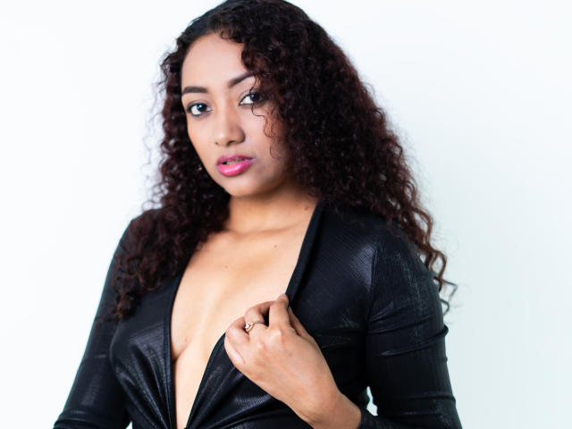 LaurenHotestx, avec ses cheveux bruns bouclés, son rouge à lèvres rose et sa peau foncée, porte un haut noir texturé avec un décolleté en V profond, qui révèle sa poitrine, et tient le haut avec sa main droite..