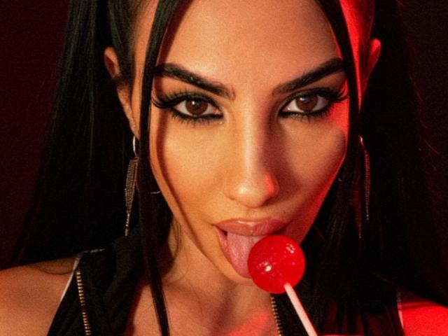 BrunetteBabe69 with long black hair, thick eyelashes, and hoop earrings, sticking a red lollipop into her slightly open mouth, tongue out, with red lighting on her face.
