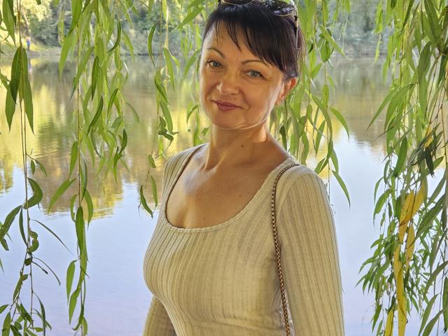 AuroraRosco with medium tits, wearing a ribbed, white, long-sleeve top, standing under green willow leaves, smiling, and having brown hair with a slight updo.
